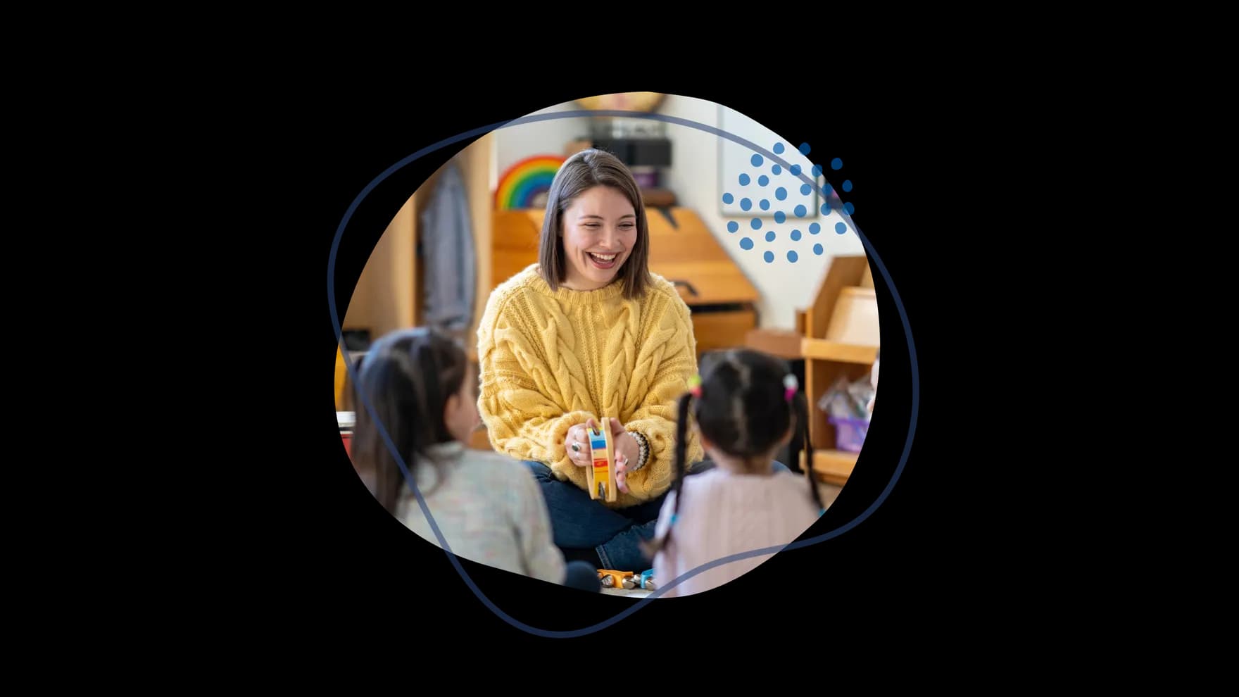 Eine Frau mit gelbem Pullover lächelt und spricht mit zwei Kindern, während sie ein buntes Spielzeug in einem Raum hält.